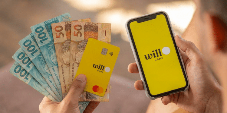 Mãos segurando cartão amarelo Will Bank Mastercard, notas de R$50 e R$100, e celular com app Will Bank aberto