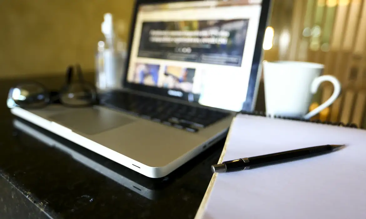Notebook aberto ao lado de caderno caneta e xícara representando o ambiente de home office do servidor do INSS
