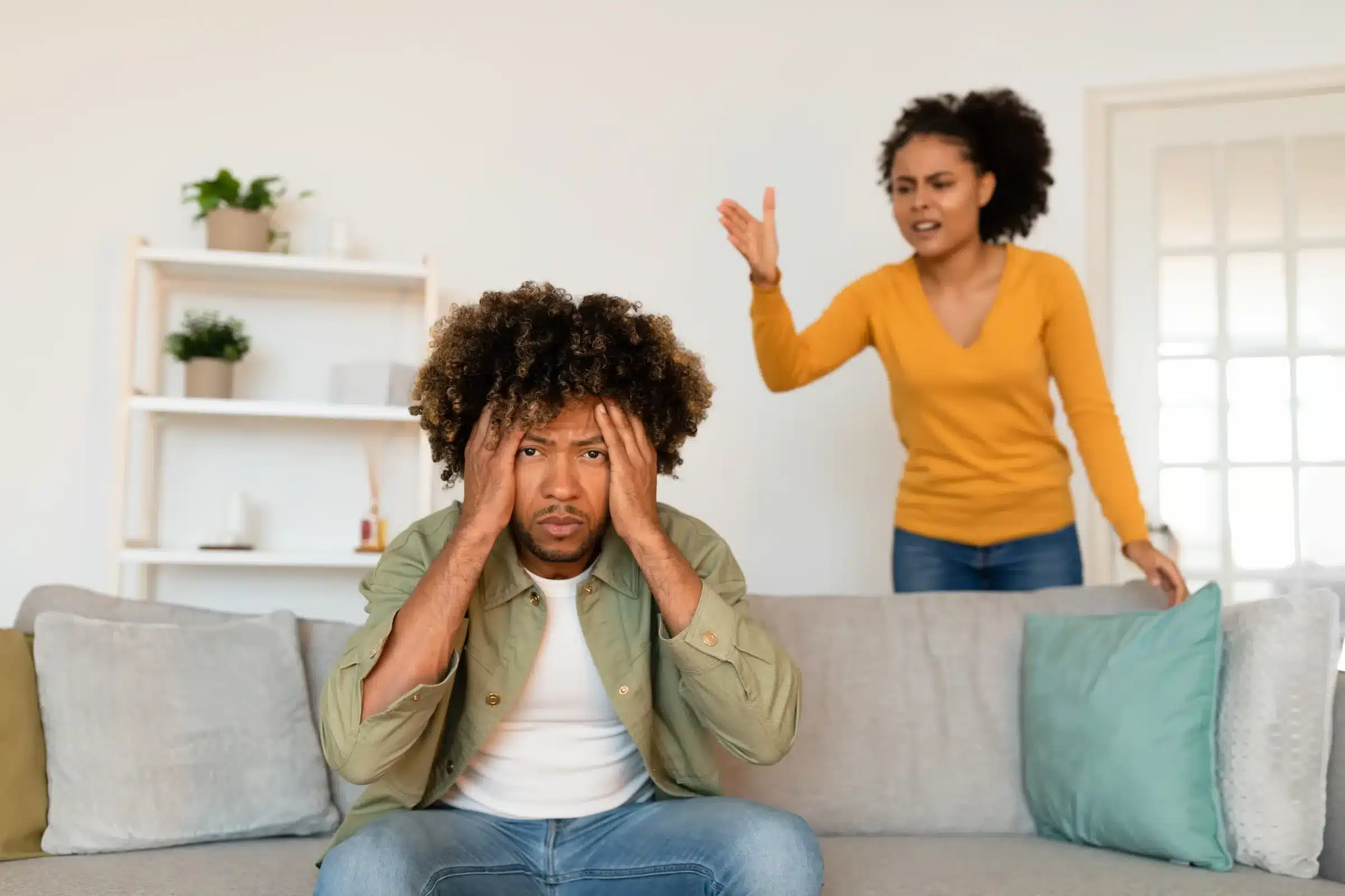 Homem sentado em sofá com as mãos na cabeça, em conflito com mulher de pé ao fundo, em um cenário de discussão, simbolizando relacionamentos tóxicos.