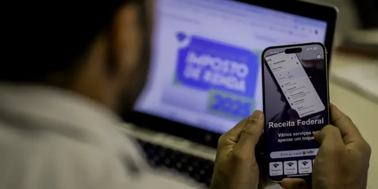 Homem consulta no celular o aplicativo da Receita Federal enquanto a página do Imposto de Renda 2026 está aberta no notebook ao fundo.