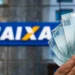 Notas de 100 reais sendo seguradas diante de uma agência da Caixa Econômica Federal, com destaque para a nova liberação de R$ 6.220.