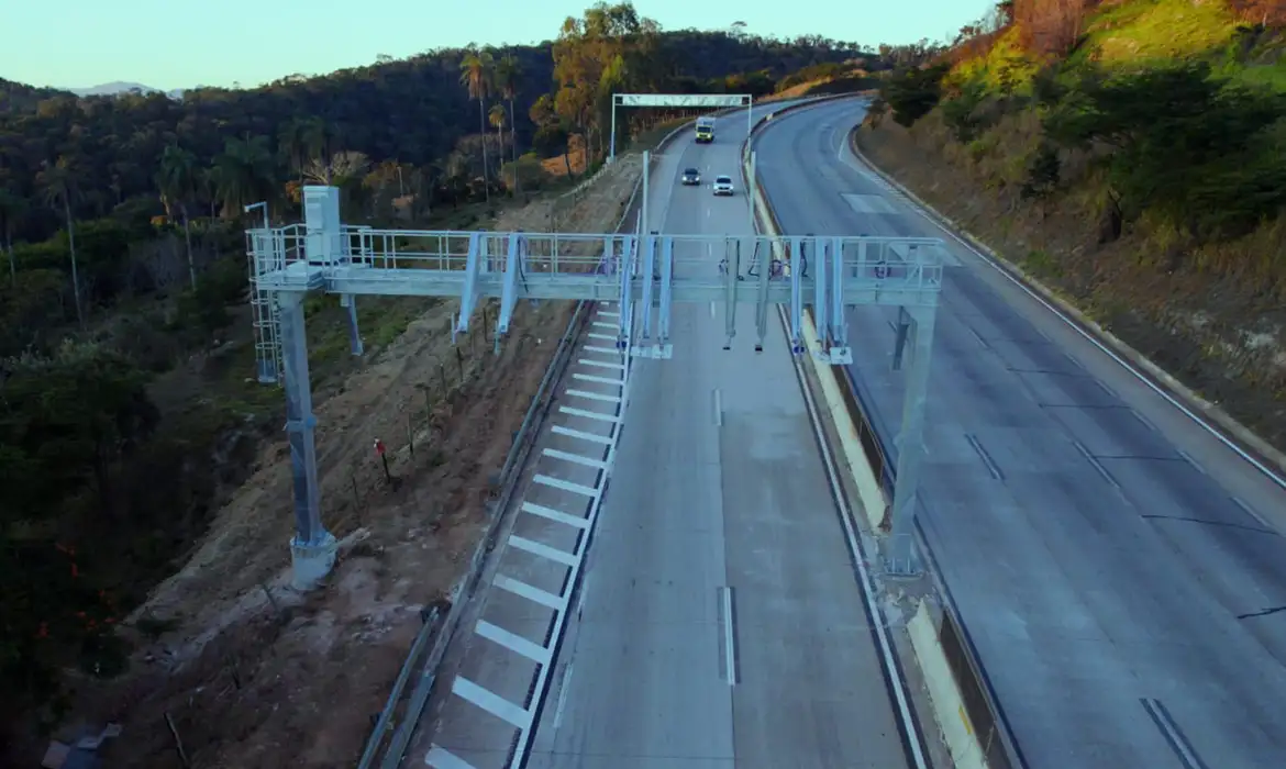 Vista aérea de pórtico de pedágio eletrônico free flow instalado sobre rodovia em área de mata, com veículos trafegando livremente sem cabines de cobrança