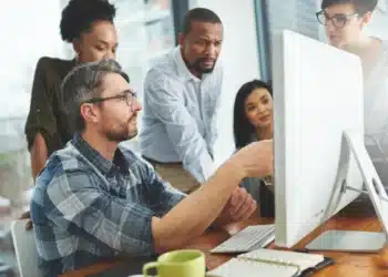 Equipe discutindo em escritório moderno, focados em tarefa na tela do computador.