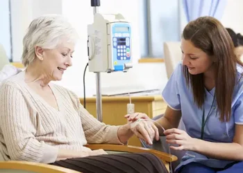 Paciente idosa sorri durante sessão de quimioterapia acompanhada por enfermeira jovem em ambiente hospitalar