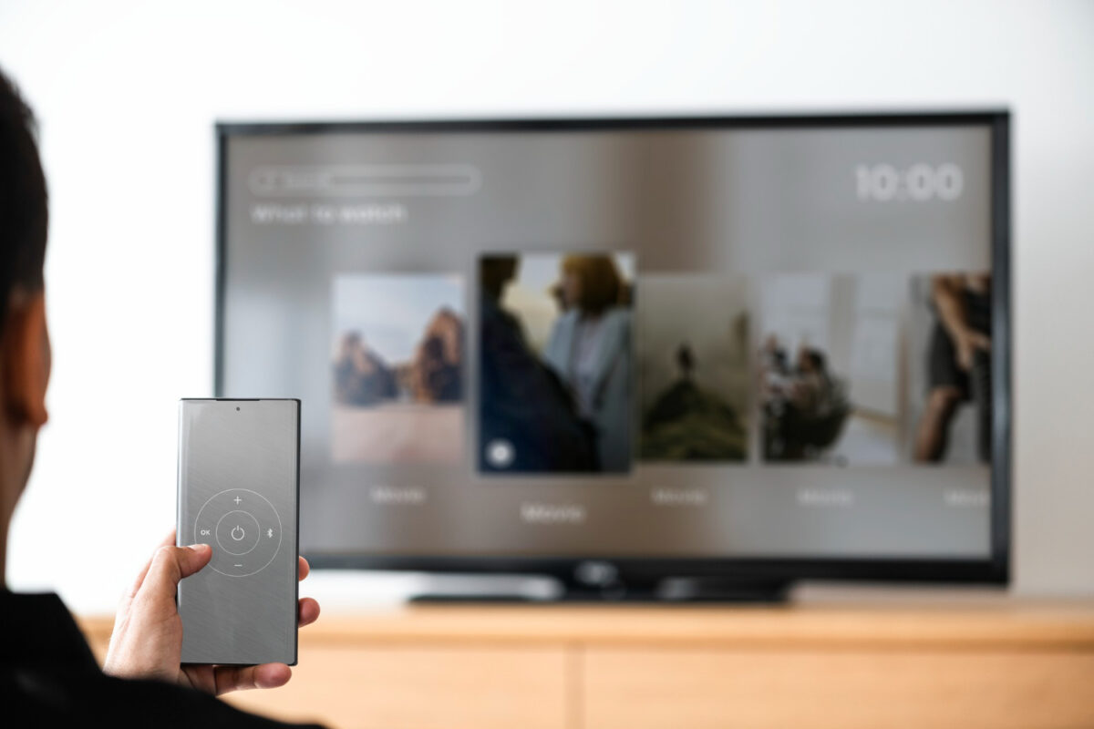 Pessoa segurando controle remoto sem botões em frente a smart TV exibindo opções de conteúdo na tela inicial