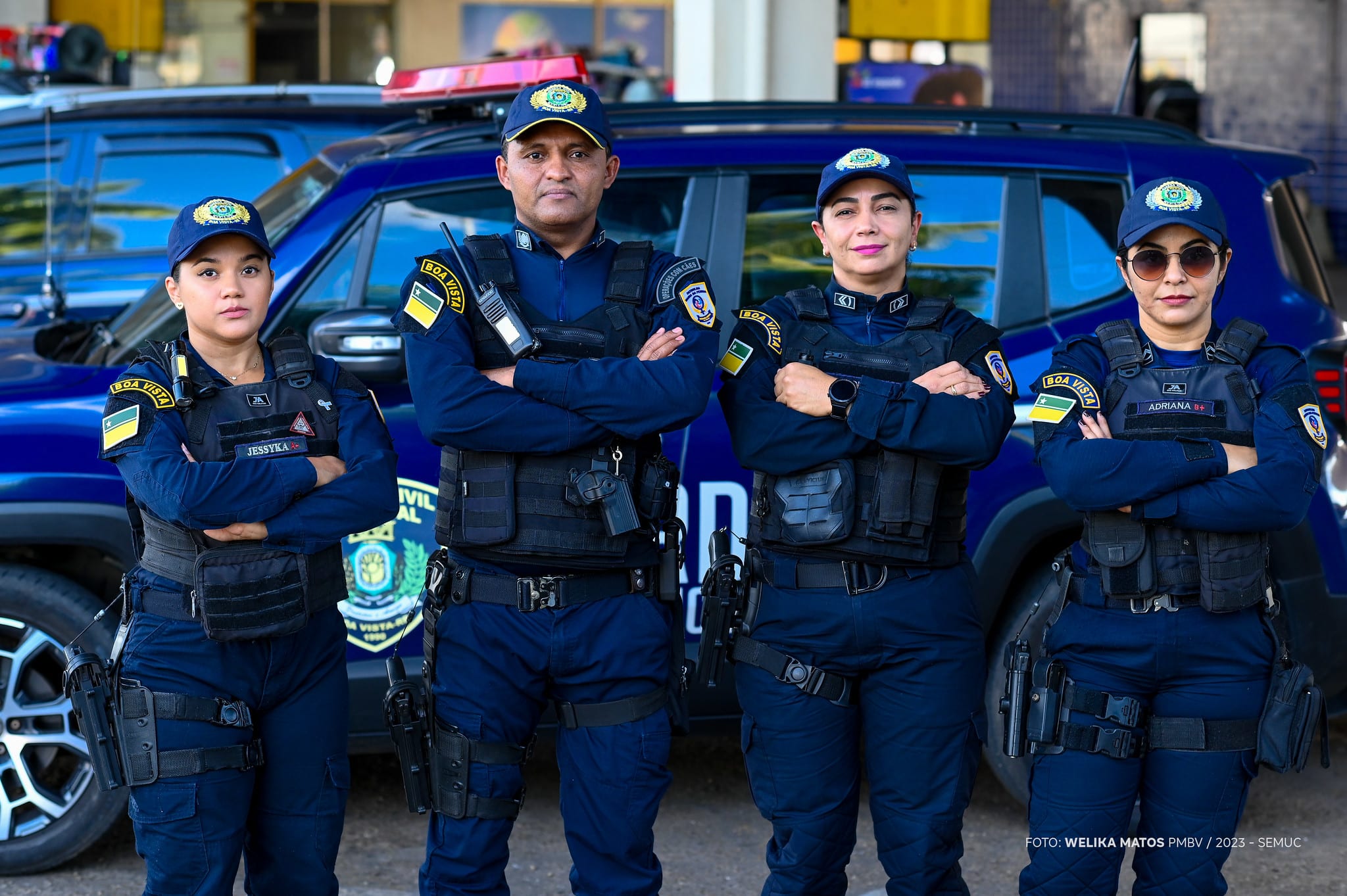 Quatro agentes da GCM de Boa Vista fardados de azul escuro, com coletes táticos, em frente a viatura azul