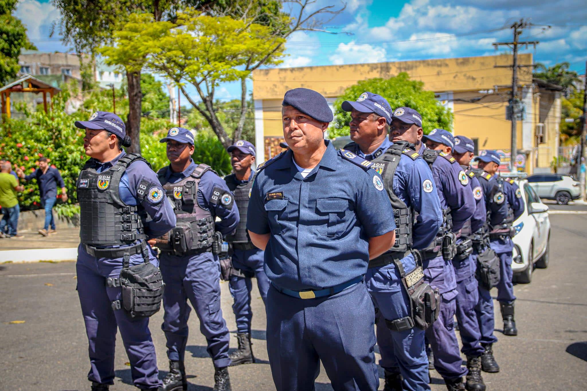 Guardas civis municipais em fila uniformizados com fardamento azul-marinho, coletes táticos e bonés em área urbana.