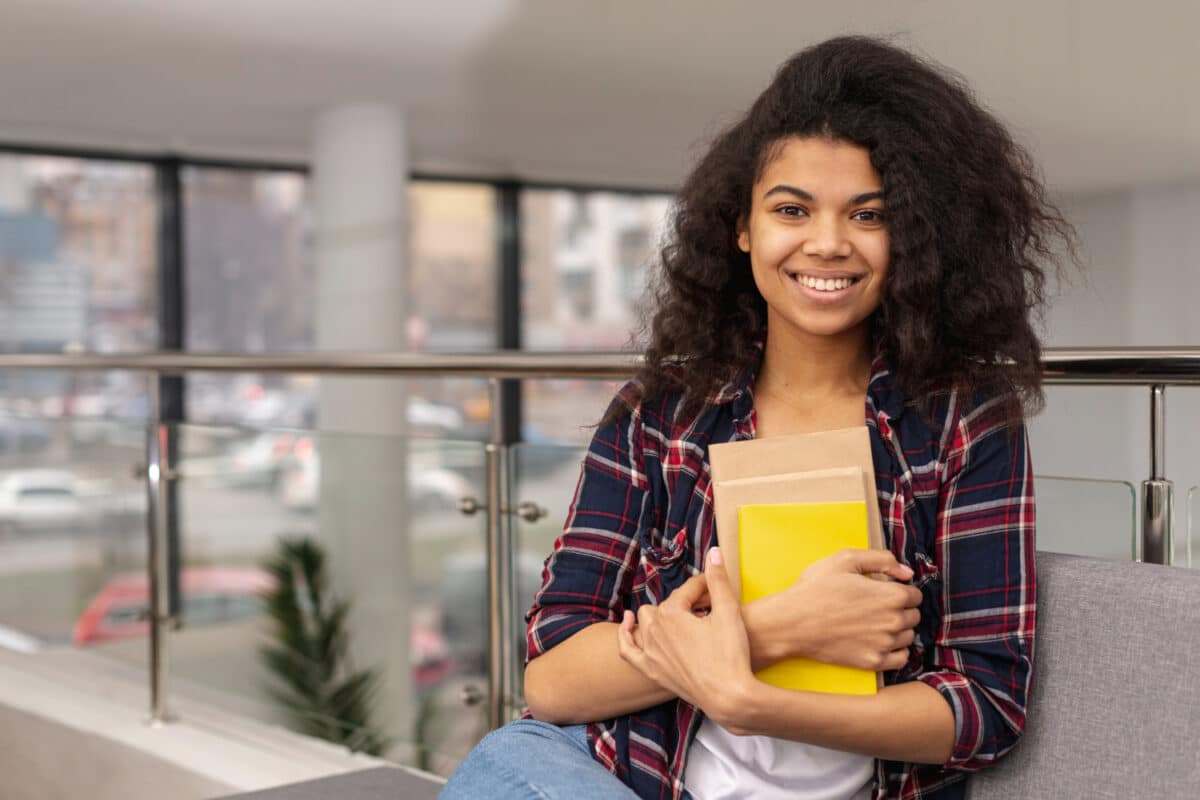 Jovem Aprendiz Correios benefícios - jovem negra sorrindo segurando livros representando as vantagens do programa