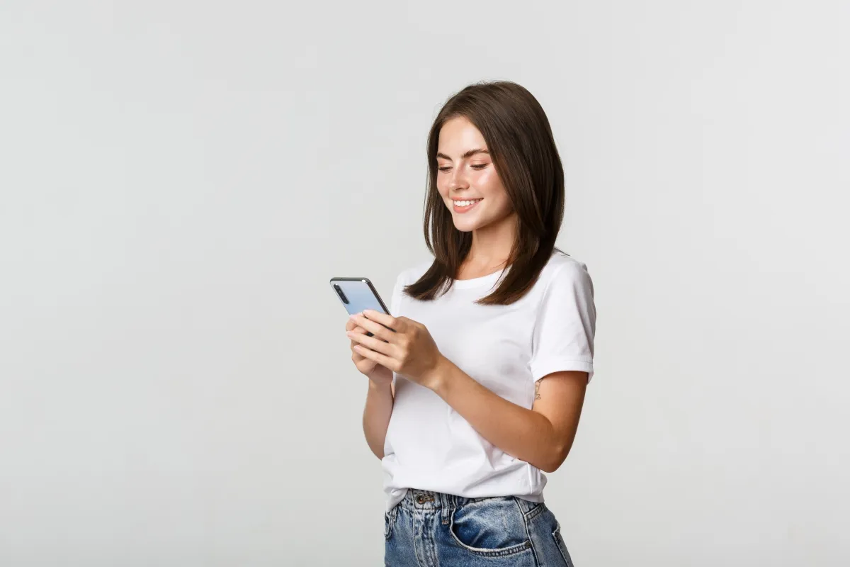 Mulher sorrindo e usando o celular, com fundo claro.