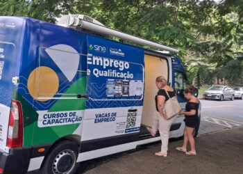 Van itinerante azul do SINE Casa do Trabalhador estacionada em área arborizada com duas mulheres no atendimento