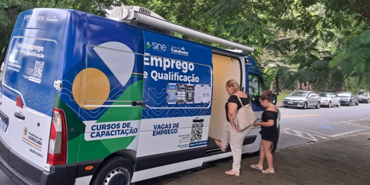 Van itinerante azul do SINE Casa do Trabalhador estacionada em área arborizada com duas mulheres no atendimento