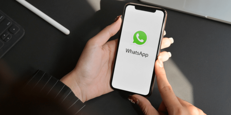 Mãos femininas com unhas vermelhas segurando iPhone exibindo logo do WhatsApp em ambiente de escritório