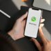 Mãos femininas com unhas vermelhas segurando iPhone exibindo logo do WhatsApp em ambiente de escritório