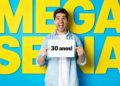 Homem jovem sorrindo segura placa "30 anos!" com fundo azul e letras amarelas "MEGA SENA".