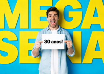 Homem jovem sorrindo segura placa "30 anos!" com fundo azul e letras amarelas "MEGA SENA".