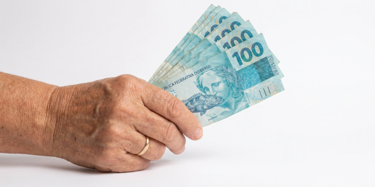Mão idosa com aliança segura notas de 100 reais em 2026.