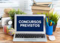 Notebook sobre mesa de madeira exibindo "CONCURSOS PREVISTOS" em tela azul, cercado por livros, câmera e plantas.