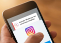 Mão segurando smartphone com notificação do Instagram sobre mudanças nas conversas protegidas e logo do app.