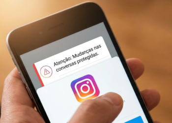 Mão segurando smartphone com notificação do Instagram sobre mudanças nas conversas protegidas e logo do app.