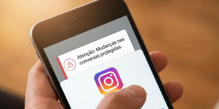 Mão segurando smartphone com notificação do Instagram sobre mudanças nas conversas protegidas e logo do app.