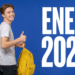 Jovem com camiseta cinza e mochila amarela faz sinal de positivo ao lado do texto ENEM 2026 sobre fundo azul.