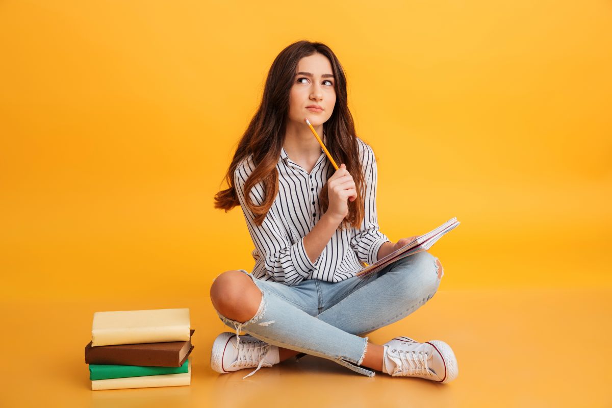 Estudante pensativa com caderno e lápis em fundo laranja.