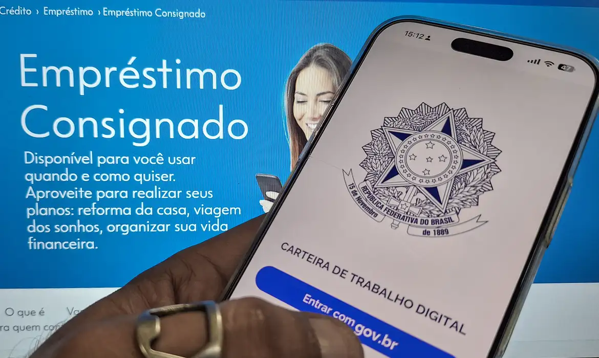 Smartphone com o aplicativo Carteira de Trabalho Digital aberto e, ao fundo, monitor exibindo página da Caixa sobre empréstimo consignado.