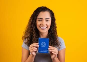 Jovem sorridente exibe Carteira de Trabalho brasileira em fundo amarelo.