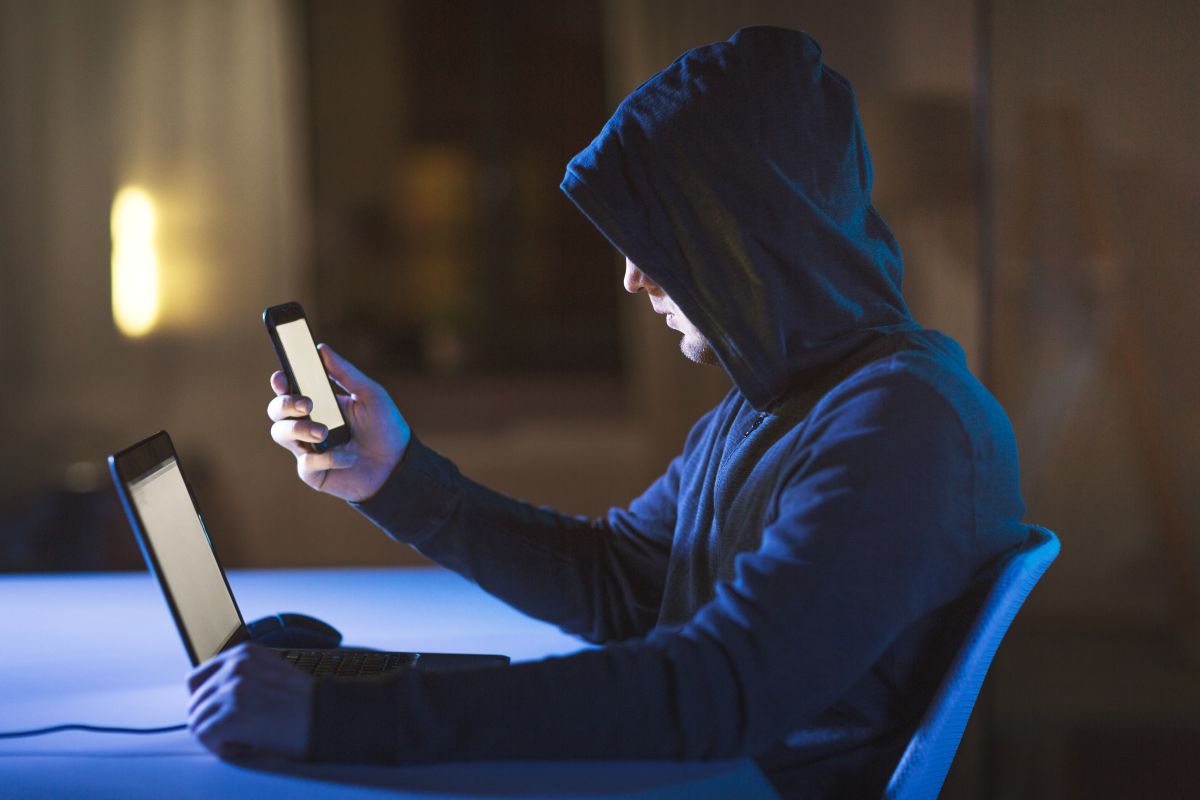 Cibercriminoso encapuzado analisa celular e laptop em sala escura.