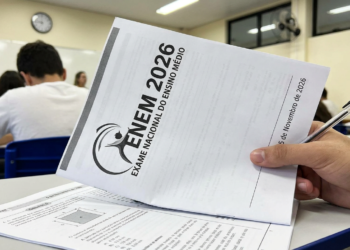 Mãos seguram caderno de provas do ENEM 2026 com caneta preta em sala de aula com carteiras azuis ao fundo.