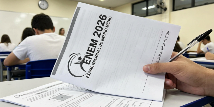 Mãos seguram caderno de provas do ENEM 2026 com caneta preta em sala de aula com carteiras azuis ao fundo.