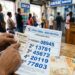 Mão segura bilhete da Loteria Federal e resultado oficial do concurso 6070 em agência da Caixa com notas de dinheiro