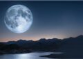 Lua cheia imensa brilha sobre lago tranquilo com reflexo prateado, cercado por montanhas escuras ao entardecer.