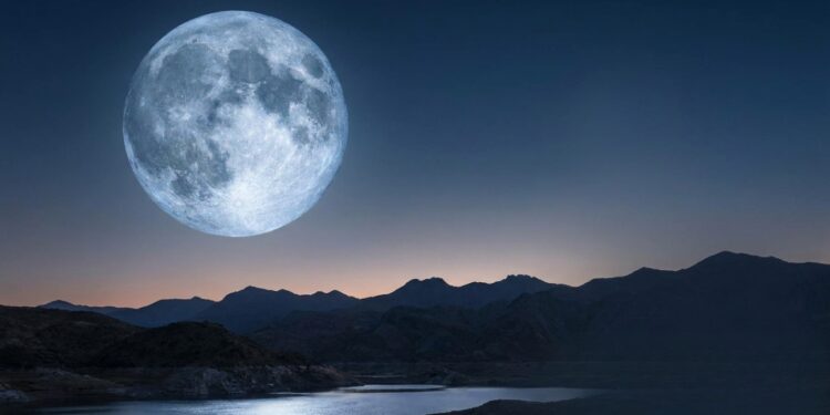 Lua cheia imensa brilha sobre lago tranquilo com reflexo prateado, cercado por montanhas escuras ao entardecer.