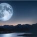 Lua cheia imensa brilha sobre lago tranquilo com reflexo prateado, cercado por montanhas escuras ao entardecer.