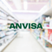 Nome "ANVISA" em corredor de farmácia com prateleiras de medicamentos.