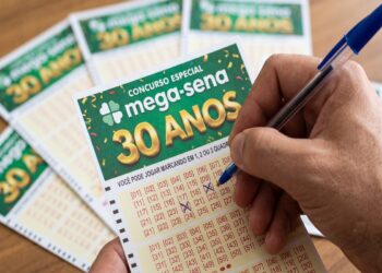 Mão segurando caneta azul marca números no volante verde do concurso especial Mega-Sena 30 Anos