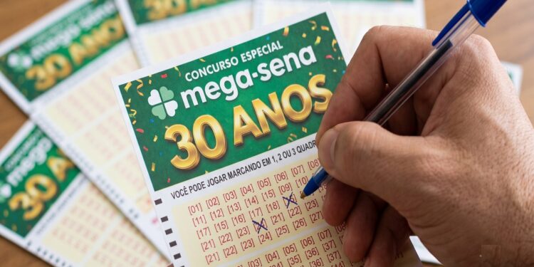 Mão segurando caneta azul marca números no volante verde do concurso especial Mega-Sena 30 Anos