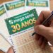 Mão segurando caneta azul marca números no volante verde do concurso especial Mega-Sena 30 Anos
