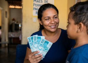Mulher sorridente segurando notas de R$100 reais ao lado de menino, parede amarela com cartaz ao fundo.
