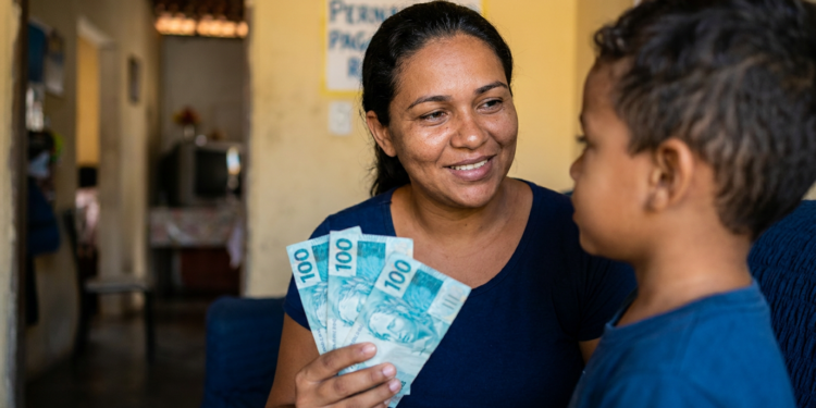 Mulher sorridente segurando notas de R$100 reais ao lado de menino, parede amarela com cartaz ao fundo.