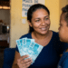 Mulher sorridente segurando notas de R$100 reais ao lado de menino, parede amarela com cartaz ao fundo.