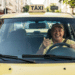Taxista sorridente em táxi amarelo nas ruas.