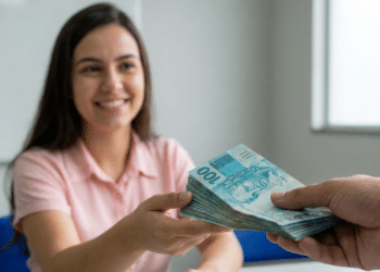 Mulher jovem de camisa rosa sorri ao receber maço de notas de R$ 100 de outra pessoa em ambiente de escritório.