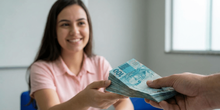 Mulher jovem de camisa rosa sorri ao receber maço de notas de R$ 100 de outra pessoa em ambiente de escritório.