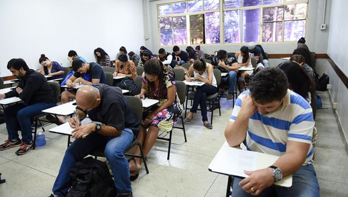 Grupo de jovens e adultos sentados escrevendo em carteiras escolares durante aplicação de prova.