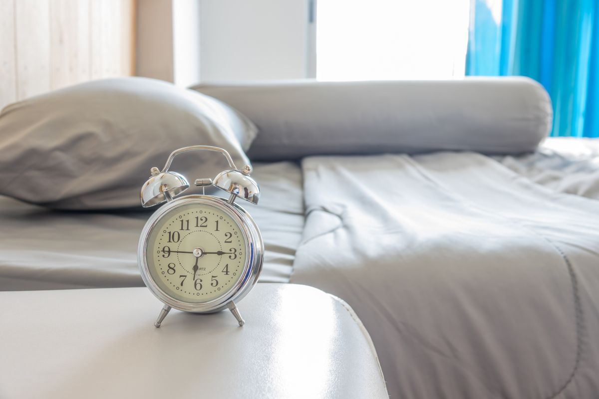 Despertador em destaque encima de uma mesa de cabeceira e cama no fundo.