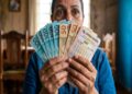 Mulher madura em camisa jeans exibe bilhete da Federal 6067 e notas de R$ 50 e R$ 100 em leque cobrindo parte do rosto