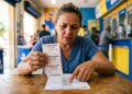 Mulher segura bilhetes da Loteria Federal sorteio 6068 em balcão de lotérica, comparando números com lista de resultados