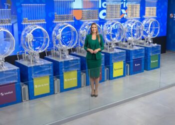 Apresentadora de blazer verde no Espaço da Sorte, rodeada por globos das loterias Lotofácil, Dia de Sorte e Quina